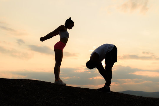 Silhouettes of fit mixed race couple stretching together on the rocky mountains background. Sport fitness concept