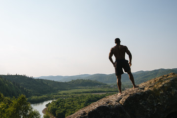 Silhouette of african american athletic man with naked torso posing on the rock. Green mountain...