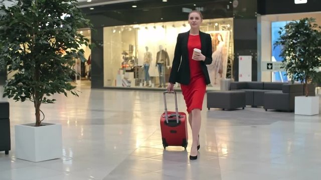 Beautiful young businesswoman in red dress pulling red suitcase across duty free zone at airport, slow motion shot on Sony NEX 700