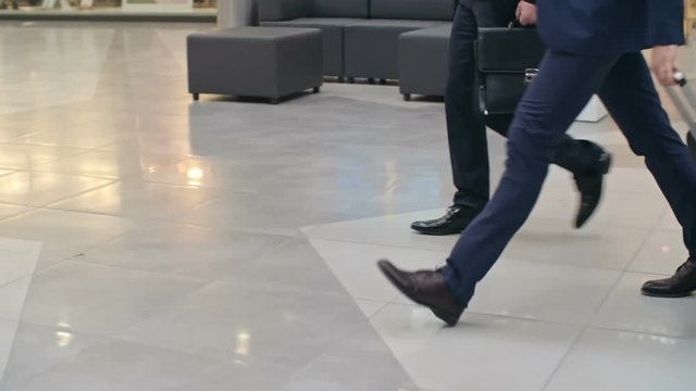 Legs of two businessmen walking on tile flooring across terminal at airport, one of them pulling suitcase; tracking slow motion shot on Sony NEX 700