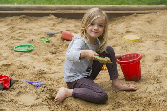 Little blonde girl is playing in sandbox with plastic toy tools