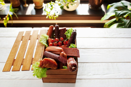Assortment of sausages in little wooden box on white wooden table. Open box with variety of sausage. Copyspace, blured background.