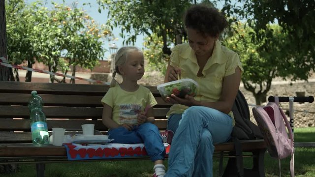 Mamma con bambina dai capelli biondi sedute a mangiare su una panchina a Roma, Italia, Europa