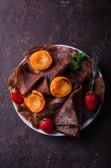Stack of chocolate crepes decorated with strawberry and grilled apricots on dark slate background. Selective focus. Top view. Background with place for some text