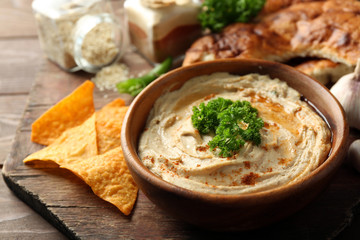 Wooden bowl of tasty hummus with chips and parsley on table
