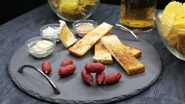 A glass with beer and snacks on a plate on a dark table. A plate of sausages , toast and sauce to beer. View of beer and snacks rotation 360 close up