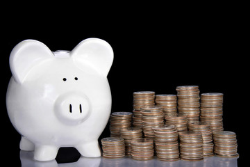 white piggy bank on a black background with stacks of quarters building up beside it reflecting on table.