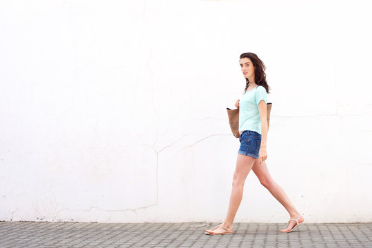 Smiling young woman with bag walking by white wall