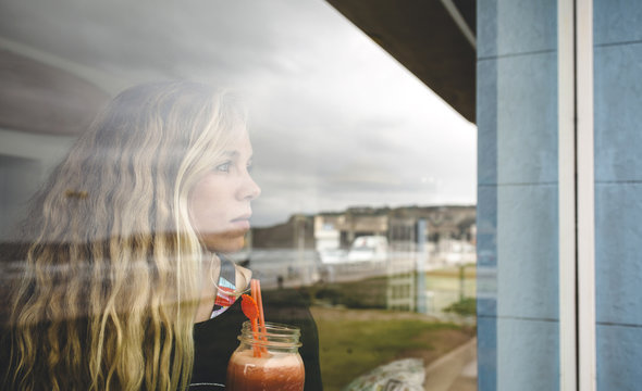 Portrait of curly blonde woman looking through the cafe window while holding a juice in her hands