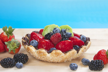 Fresh fruit tart with flower shaped pie crust sprinkled with sugar crystals coated with chocolate filled with custard topped with fresh strawberries, blue berries, boysenberries and kiwi on wood table