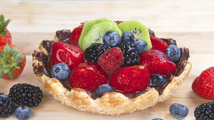 Fresh fruit tart with flower shaped pie crust sprinkled with sugar crystals coated with chocolate filled with custard topped with fresh strawberries, blue berries, boysenberries and kiwis