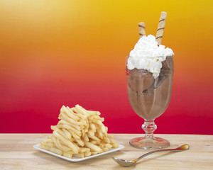 Chocolate ice cream in glass cup with whipped cream and square white plate of stacked french fries on a light wood table. Food fad or new trend. Sweet and salty. vibrant pink and yellow background.