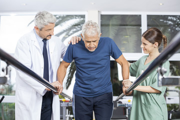 Physiotherapists Motivating Senior Patient To Walk Between Paral