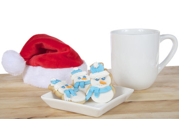 Three snowmen sugar cookies with royal icing frosting scarfs and hats on a square white plate on light wood table next to cup of coffee and miniature santa hat white background