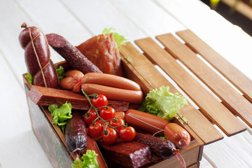 Sausage assortment in wooden box. Box with different kinds of sausages decorated with cherry tomato on white wooden background.