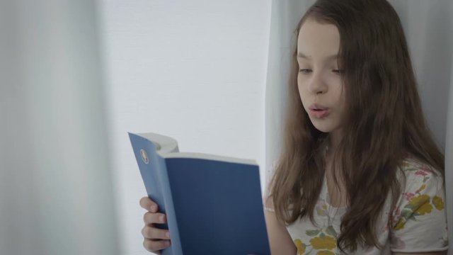 Beautiful little girl reading an interesting book by window.