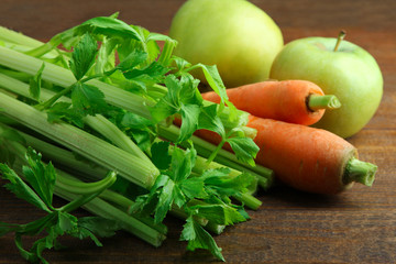 Fresh green celery with carrots and apples on a brown wooden background