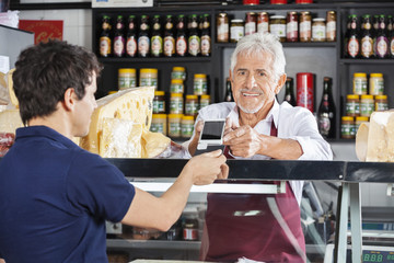 Salesman Accepting Mobile Payment From Customer In Cheese Shop