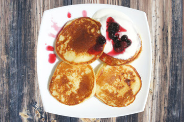 Pancakes with sour cream and berries in a on a square white plate. The cheesecakes on the plate drizzled with jam and sour cream. Hot cakes for Breakfast in white plate on wooden texture, top view