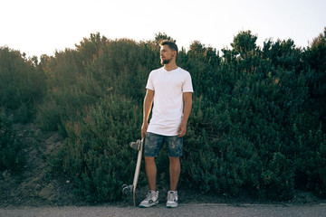 Young male wearing a blank white t-shirt and shorts is looking aside. A skater with a board is standing on a bush wall background.