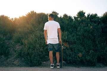 A man wearing a blank white t-shirt and shorts is standing with his back to the camera. A skater boy with a skateboard is looking aside on a bushy background. © JKstock