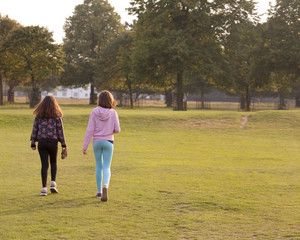 Fototapeta premium Heading Home. Two young sisters walk together towards the exit of the park. The sun is setting and the low afternoon light is outlining their figures.