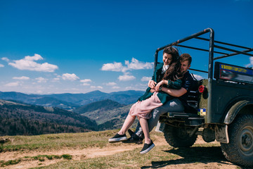 Side view of a smiling woman embracing man against  offroad old jeep and mountains. Woman in dress. Man show on landscape. Blue sky sunny day. Amazing travel