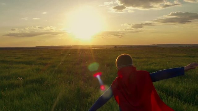 superhero boy running on the green field at sunset