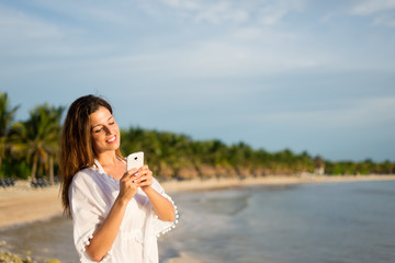 Happy woman on tropical summer vacation texting on smartphone towards the sea. Female tourist messaging on travel to Riviera Maya, Mexico.