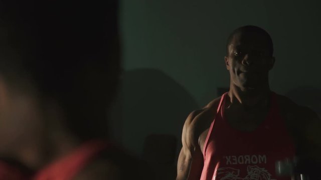 Black muscular african-american bodybuilder training at the gym with dumbbells and looks on mirror reflection on dark background 