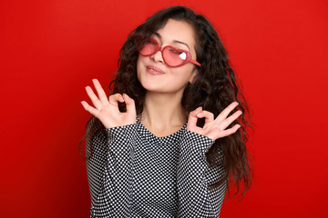 young woman beautiful portrait show okay sign, posing on red background, long curly hair, sunglasses in heart shape, glamour concept