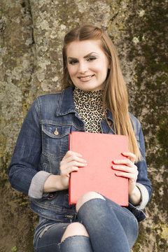 Eine junge braunhaarige Frau in Jeans sitzt in einem Park und liest in einem roten Buch.