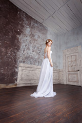 Fototapeta na wymiar Beautiful young bride in a long wedding dress is standing in an empty room. White flowers in her red hair. She is pale, elate and tender.