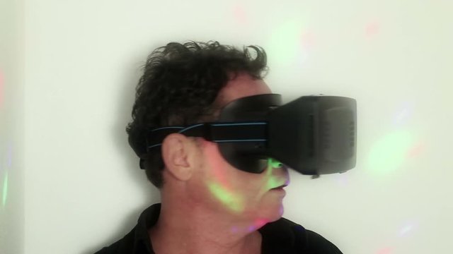Medium CU of a man reacting to on-screen action inside his virtual reality (VR) headset. At one point he looks up towards some illusory high point (*more action and external lighting effects)