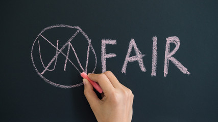 Hand using chalk changing the word unfair to fair by crossing the word un on the blackboard,...