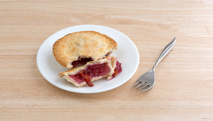 Side view of a small cherry pie on a plate with a fork atop a wood table top