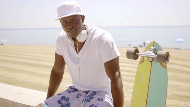 Black skater next to his board and sandy beach checks his phone while wearing white shirt and headset around his neck