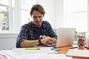 Man Sitting At Desk In Home Office Looking At Smart Watch