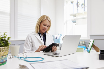 Obraz na płótnie Canvas Female Doctor Sitting At Desk Using Digital Tablet In Office