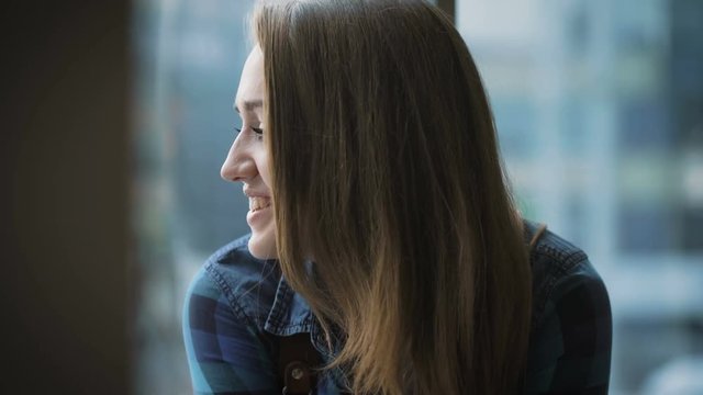 Portrait of attractive girl with long hair. Student sitting near the window and friendly talks with someone.