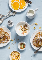 Whole wheat pancakes, greek yogurt with homemade granola, orange slices, nuts, corn flakes, on a blue stone background.  Breakfast table.