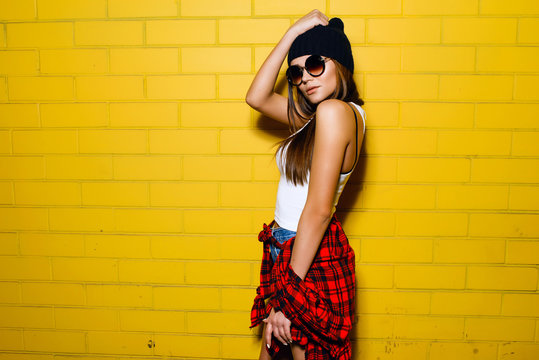 Beautiful young sexy hipster girl posing and smiling near urban yellow wall background in sunglasses, red plaid shirt, shorts, hat, sneakers.