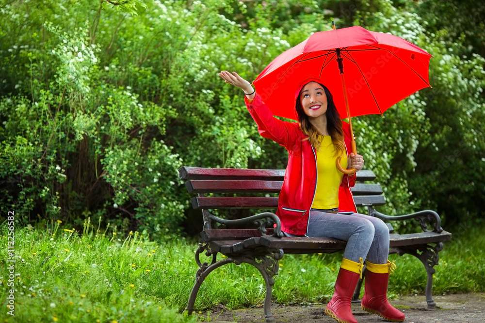 Wall mural Young woman sitting on a park bench with red umbrella checking is it raining.