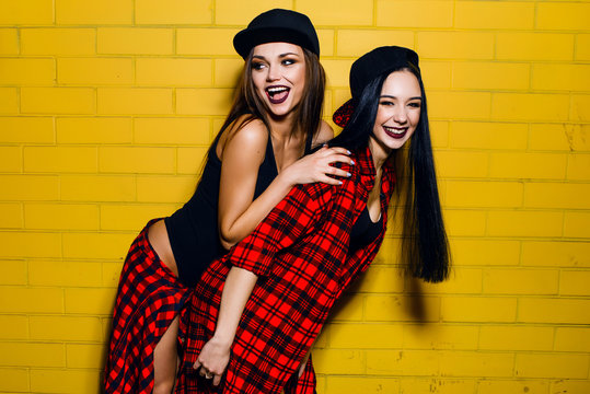 Two young happy hipster girls having fun, smiling, laughing, jumping, walking  near urban yellow wall background, summer relax concept