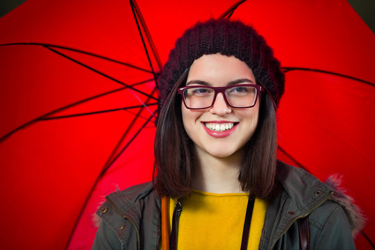 Portrait of smiling hipster girl holding a red umbrella and looking at camera. She is wearing eyeglasses and a hat. Copy space