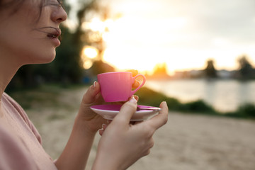 female's hands keep cup of coffee near lake or river;