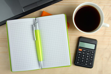 green notebook on a brown wooden background with a pen and a calculator near the laptop with a strong Cup of coffee