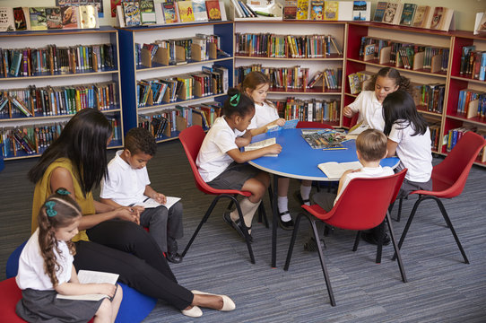 Elementary School Pupils Reading In Library With Teacher