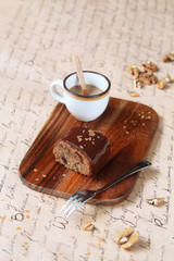 Piece of Date Cake with Walnuts and Chocolate Mirror Glaze on a wooden cutting board, and a cup of coffee, on a light beige background.