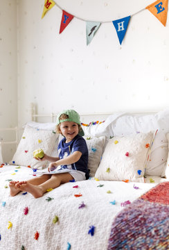 Portrait of smiling little boy sitting on the couch eating an apple and playing with a tablet